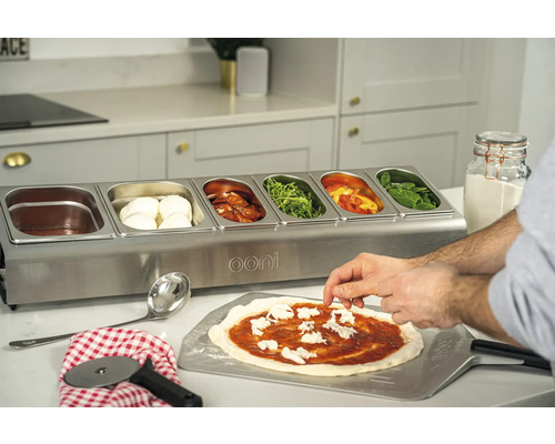 Ooni bereidingsstation met ingrediënten en persoon die een pizza bereidt.
