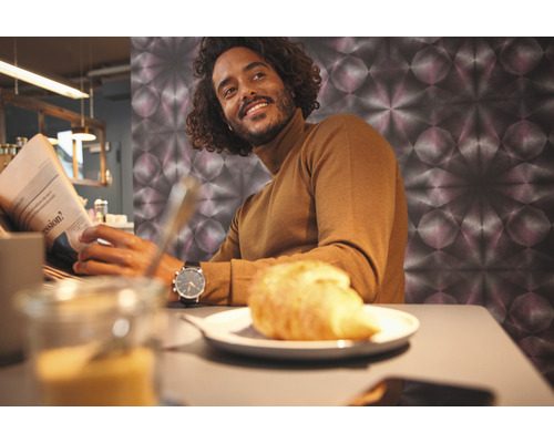 Een man leest de krant tijdens het ontbijt voor een muur met grafisch patroon.