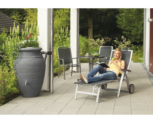 Scene met een vrouw die ontspant op een ligstoel, een regenton in de vorm van een amfora en tuinmeubelen op een terras.