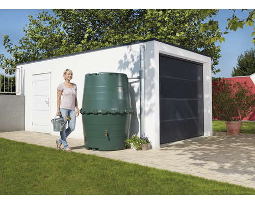 Een vrouw staat met een gieter naast een regenton voor een garage.