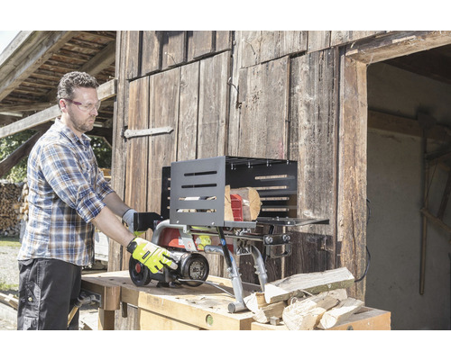 Een man bedient een houtklover voor een houten wand. De houtklover staat op een werkbank en er liggen houtblokken naast hem.