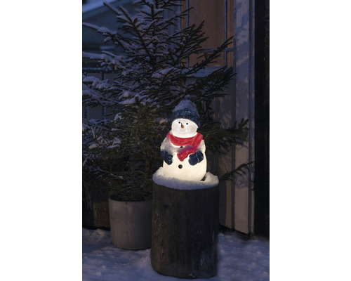 Verlichte sneeuwpop met muts en sjaal op een boomstronk, omringd door een besneeuwde kerstboom.