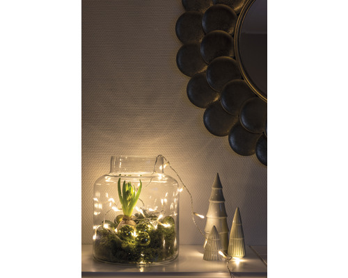 Decoratieve glazen pot met hyacint, mos, ballen en lichtsnoer, naast decoratieve keramische bomen en een ronde spiegel