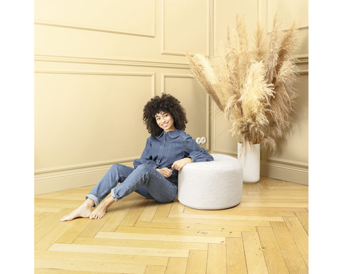 Vrouw zit op een ronde, witte kruk in een kamer met een houten vloer
