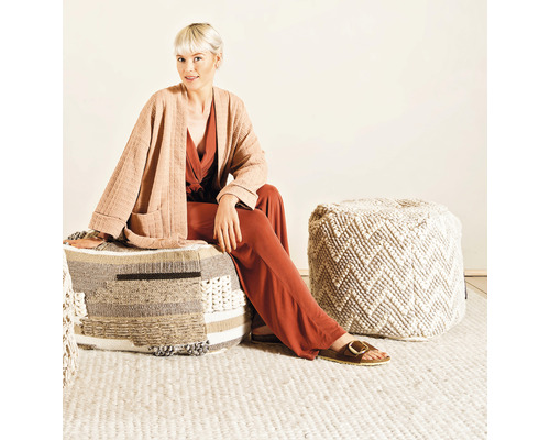 Vrouw zit op een vierkante poef gemaakt van verschillende materialen in een heldere kamer.