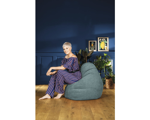 Vrouw zit in een kamer op een zitzak, omringd door kamerplanten en afbeeldingen.