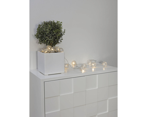Decoratieve lichtketting met bolvormige lampen op een witte commode naast een potplant.
