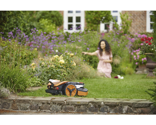 Een robotmaaier staat op het gazon voor een bloeiende tuin met een vrouw op de achtergrond.