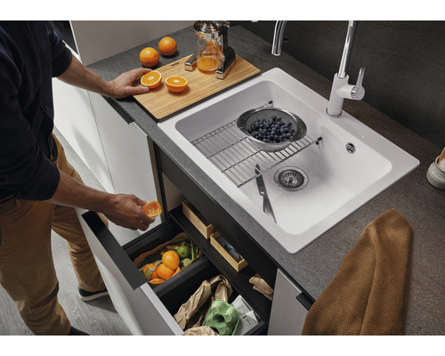 Keukenscène met spoelbak, kraan, snijplank, citruspers en afvalemmer onder de spoelbak.