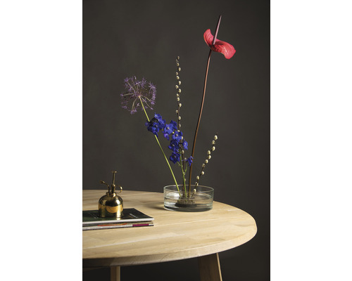 Bloemstuk met anthurium, sierui en delphinium in glazen vaas op houten tafel, met een gouden plantenspuit ernaast