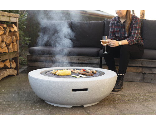 Vuurplaats met grillfunctie en bereide gerechten erop in de tuin, een vrouw zit in de buurt