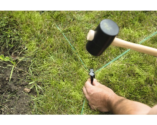 Iemand bevestigt een groen richtsnoer met een hamer en een haring in het gazon.