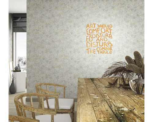 Woonkamer met houten tafel, stoelen en wanddecoratie met de tekst Kunst moet de gestoorden troosten en de comfortabelen storen.