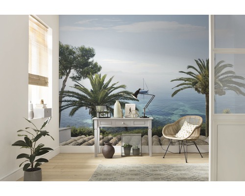 Woonkamer met fotobehang dat een kustlandschap met palmbomen en een zeilboot laat zien, ingericht met een bureau en een rotan stoel.