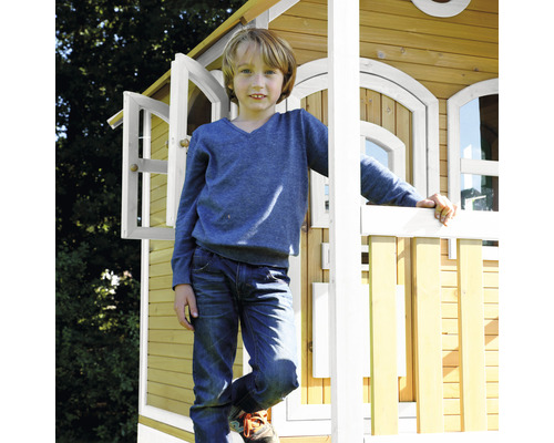 Jongen staat voor een houten speelhuis met open ramen en een hek