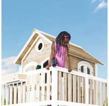 Een kind speelt op een houten speelhuis met veranda tegen een blauwe lucht.