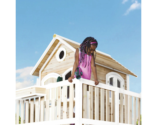 Een kind staat op een houten speelhuis met een balustrade en een klein huis met ramen onder een blauwe lucht.