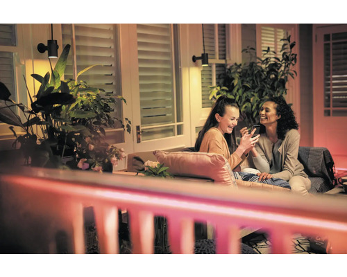 Scene met twee vrouwen die op een terras onder buitenwandlampen zitten en naar een smartphone kijken, omringd door planten.