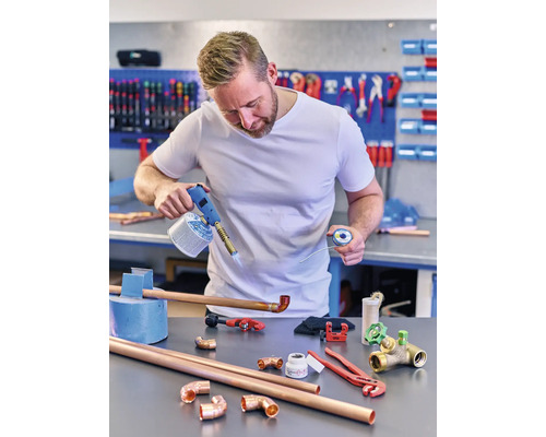 Een man soldeert koperen buizen met een gasbrander, soldeer, buizen en fittingen op een werkbank.