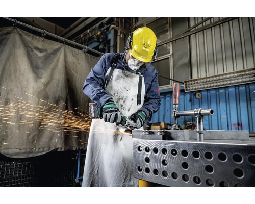 Een arbeider met veiligheidshelm, gehoorbescherming, ademhalingsmasker en schort slijpt metaal met een haakse slijper, waarbij vonken vrijkomen.