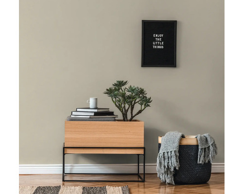 Woonkamerscène met een houten tafel, een plant, boeken, een beker, een mand met een deken en een ingelijst citaat aan de muur.