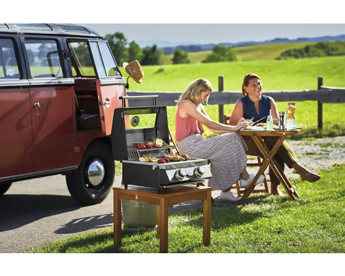 Scene met een draagbare gasbarbecue, een tafel en twee personen buiten naast een rode oldtimerbus