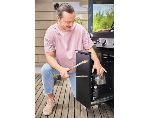 Een man knielt voor een gasbarbecue en houdt een barbecuetang in zijn hand. De deur van de onderkast is geopend en toont barbecue-accessoires.