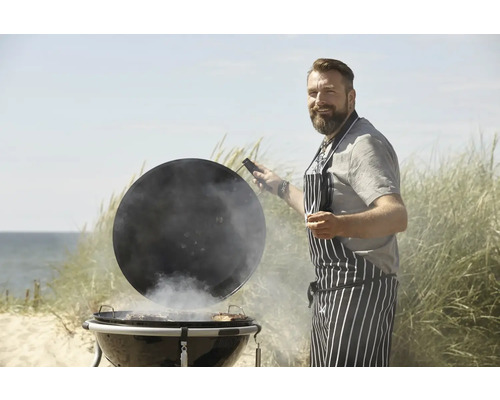 Man met grillschort opent het deksel van een kogelbarbecue op het strand