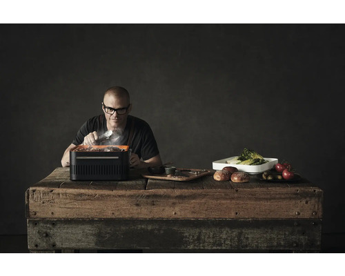 Scene met tafelgrill, barbecuevlees, salade, tomaten, broodjes en een persoon die aan het grillen is