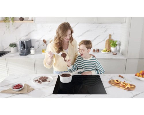 Moeder en zoon bereiden chocolade in een lichte keuken op een inductiekookplaat. Aardbeien, chocolade en perziken staan op het aanrecht.