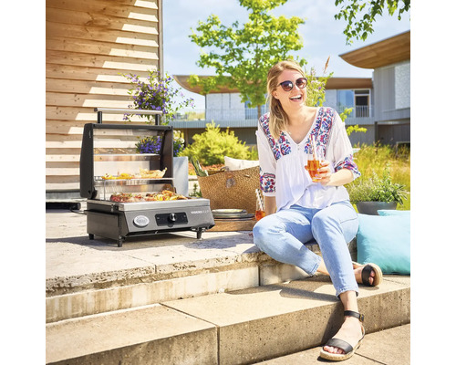 Buitenscène met een tafelbarbecue, bereide gerechten en een vrouw die van een drankje geniet