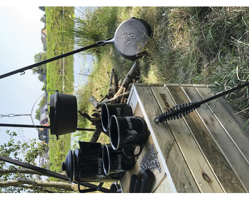 Kampvuur kookplaats met driepoot, pan, koffiepot, mokken en grillbestek op een houten tafel