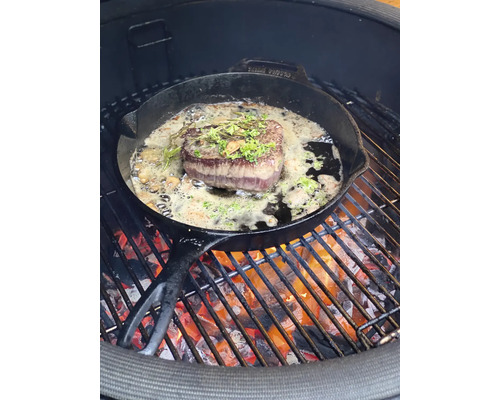 Een steak wordt bereid in een gietijzeren pan op een barbecue.