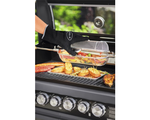 Een persoon met een barbecuehandschoen bereidt groenten in een barbecue-mand op een gasbarbecue, samen met worstjes en knoflookbrood.