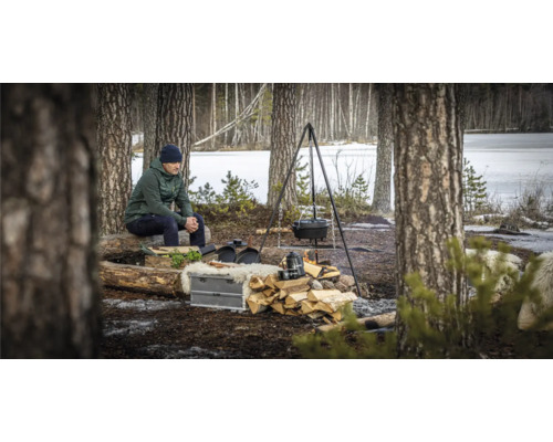 Buitenscène met driepoot, Dutch oven, stapel hout en zittende man in de natuur