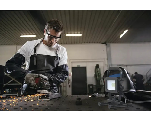 Een man met veiligheidsbril, schort en handschoenen slijpt metaal met een flex, terwijl een bouwlamp op de voorgrond staat.