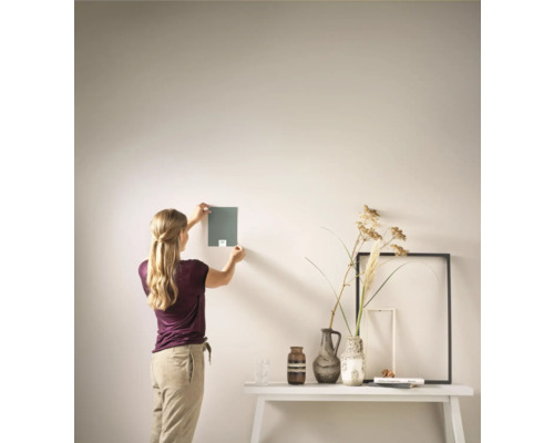 Vrouw houdt kleurstaal tegen muur boven een tafel met vazen en fotolijsten.