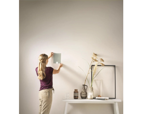 Vrouw houdt een kleurmonster tegen een muur in een lichte kamer met een tafel en decoratie.