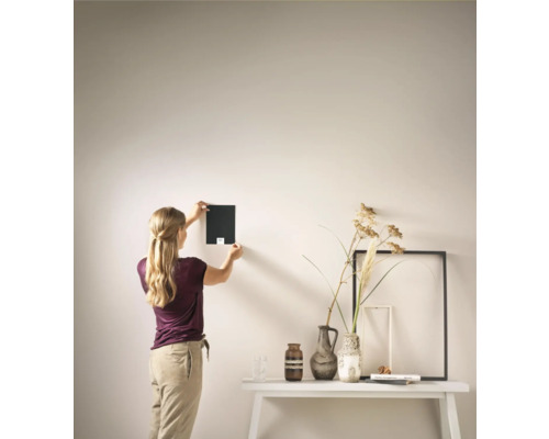 Vrouw hangt kleurstaal aan de muur boven een tafel met vazen en fotolijsten