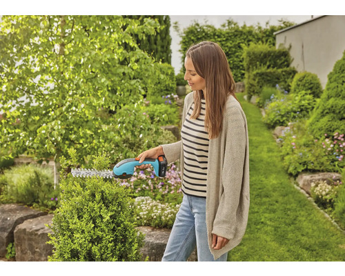 Een vrouw snoeit een heg met een accuheggenschaar in een tuin.