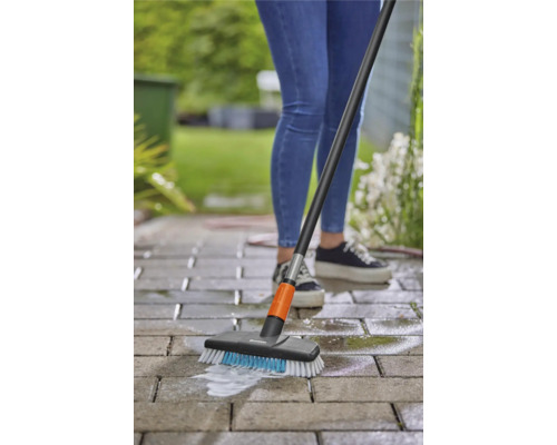 Een persoon reinigt bestrating met een borstel en steel buitenshuis.