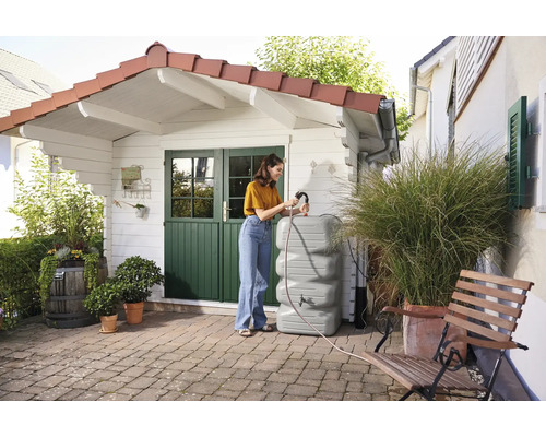 Vrouw vult een grijze regenwatertank op een terras voor een tuinhuis.