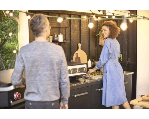 Scène van een buitenkeuken met pizzaoven, verlicht met een lichtsnoer, terwijl een stel eten bereidt