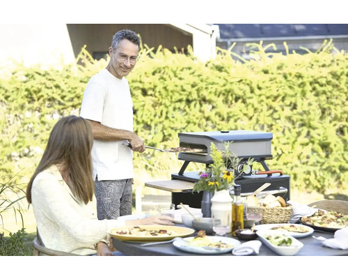 Buitenscène met een man die pizza bereidt in een pizzaoven terwijl een vrouw aan tafel zit te eten.