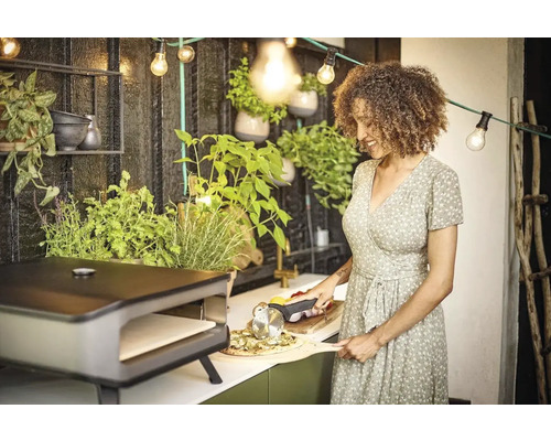 Een vrouw snijdt pizza met een pizzasnijder naast een pizzaoven op een terras.