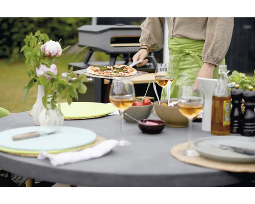 Scene van een pizza die met een pizzaschep uit een pizzaoven wordt gehaald, gedekte tuintafel met glazen en gerechten