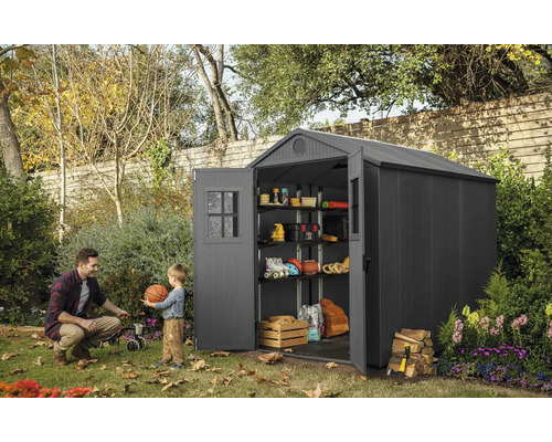 Tuinscène met een kunststof tuinhuis met open deuren, planken en opbergartikelen, een man en een kind spelen met een bal