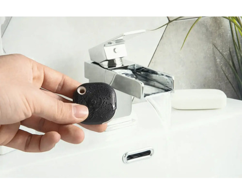 Hand houdt een zwarte, vierkante waterdetector vast voor een wastafel met kraan