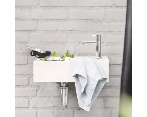 Een wastafel met kraan, handdoek, beker en plant staat voor een lichte bakstenen achtergrond.