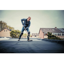 Een man veegt dakbedekking op een plat dak met een bezem.